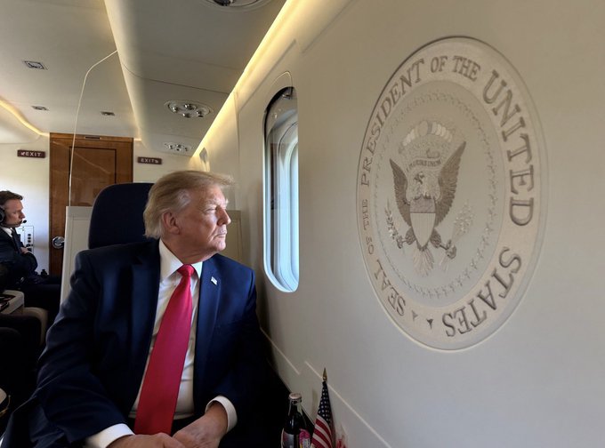 Donald Trump sits in a dark suit with a red tie inside an aircraft, looking out the window next to the large 'SEAL OF THE PRESIDENT OF THE UNITED STATES' emblem on the cabin wall.