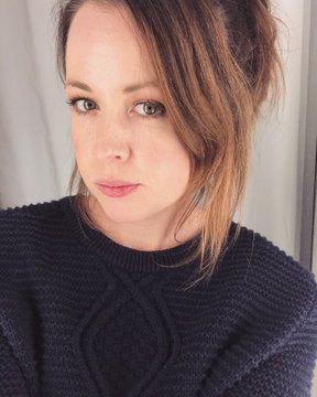 A selfie of a woman with brown hair pulled back, wearing a dark navy cable-knit sweater, looking directly at the camera with a neutral expression.