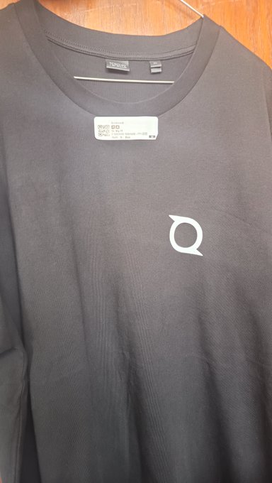 A close-up photo of a black t-shirt hanging on a hanger, featuring a small white circular logo resembling the Solana emblem on the chest area.