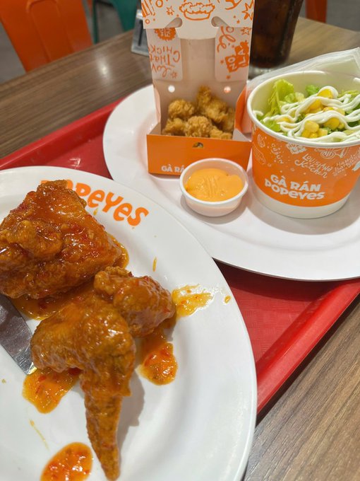 A Popeyes meal is displayed on a red tray, featuring spicy orange-glazed chicken wings on a branded plate, alongside popcorn chicken, coleslaw, and a dipping sauce.