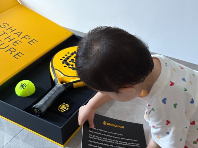 A young child in patterned pajamas looking at a yellow BNB Chain branded box containing a tennis racket, tennis ball, and a black cap, while holding a BNB Chain informational card.