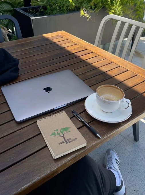 MacBook, coffee, pen, and a "Save the Planet" notebook arranged on a wooden café table—embodying a trader or investor's morning routine of consistent study and journaling, perfectly complementing the post's message about daily 1% improvements compounding over chasing big wins.