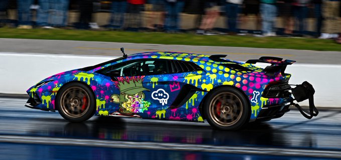 A side view of a Lamborghini on a road featuring a colorful, vibrant graffiti-style wrap with neon polka dots, drip patterns, and a cartoon character on its door panel.