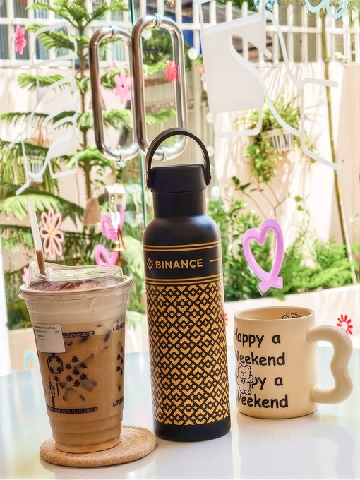 The post caption says "good monday (this mug is lying)" — a cheeky nod to the Monday blues — which matches the scene of a Binance-branded gold and black insulated water bottle next to an iced coffee and a ceramic mug that ironically declares "Happy a Weekend a py a eekend" (a playful misprint of "Happy a weekend"), all sitting on a sunlit café table surrounded by lush greenery and colorful sticker decorations.
