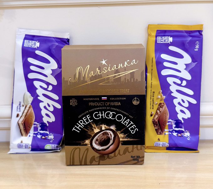A display of chocolate products featuring a 'Marsianca' Three Chocolates box (Product of Russia, 150g) flanked by two Milka chocolate bars, one purple (Milka Alpine Milk) and one yellow (Milka with hazelnuts).