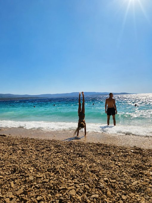 A sunny beach scene shows a person doing a handstand in the shallow turquoise water while another person stands nearby in the surf, with a rocky shoreline in the foreground and distant mountains under a clear blue sky.