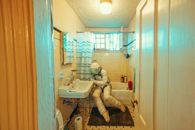 An astronaut in a full white spacesuit and helmet slumps lifelessly against a bathroom sink in a small, tiled bathroom with a shower curtain, blue ceiling, and a window above the tub.