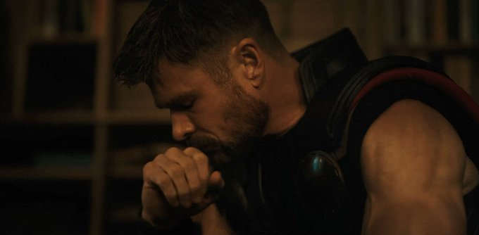 A scene featuring actor Chris Hemsworth as Thor, looking down with his hand near his mouth in a contemplative pose, wearing his dark armored suit.