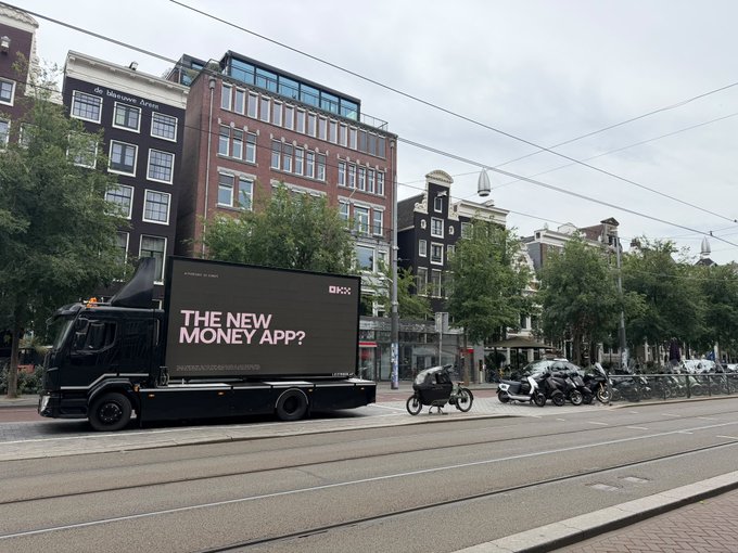 An OKX mobile billboard truck displaying "THE NEW MONEY APP?" drives down an Amsterdam street lined with classic Dutch architecture, supporting the post's message about OKX expanding its presence in Europe amid shifting crypto regulations.