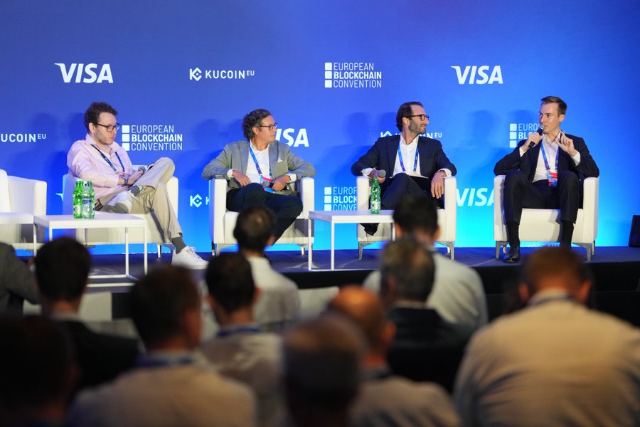 Four men in business attire sit on white chairs on a stage participating in a panel discussion at the European Blockchain Convention, with Visa and KuCoin EU branding displayed on the blue backdrop behind them.