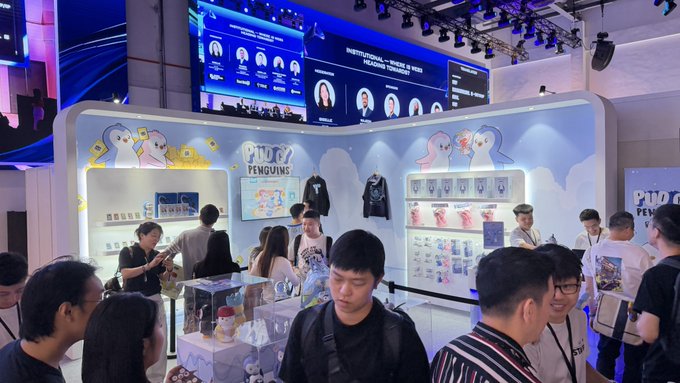 A photograph of a bustling 'PINGU PENGUINS' merchandise booth at an indoor convention, with attendees browsing products and a large digital display showing portraits overhead.
