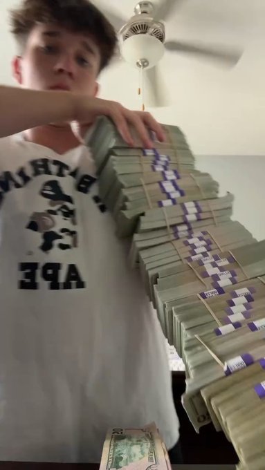 A young man wearing a backwards 'APE' graphic t-shirt stands indoors holding up a large, rubber-banded stack of banded US currency bundles, with a ceiling fan visible above.