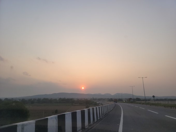 A scenic landscape captures a sunset with the sun low on the horizon over distant mountains, viewed from a curving highway road with a striped barrier wall in the foreground.
