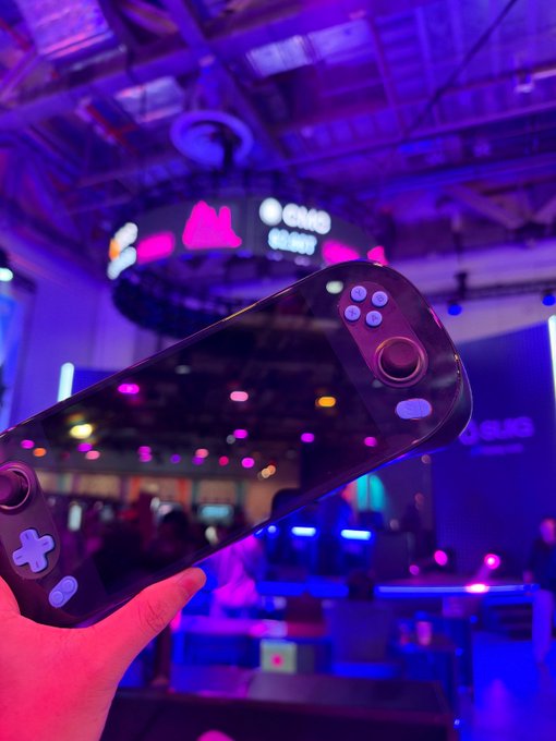 A hand holds a transparent black handheld gaming console in a vibrant purple and pink neon-lit entertainment venue, with an 'OMG' sign visible in the background.