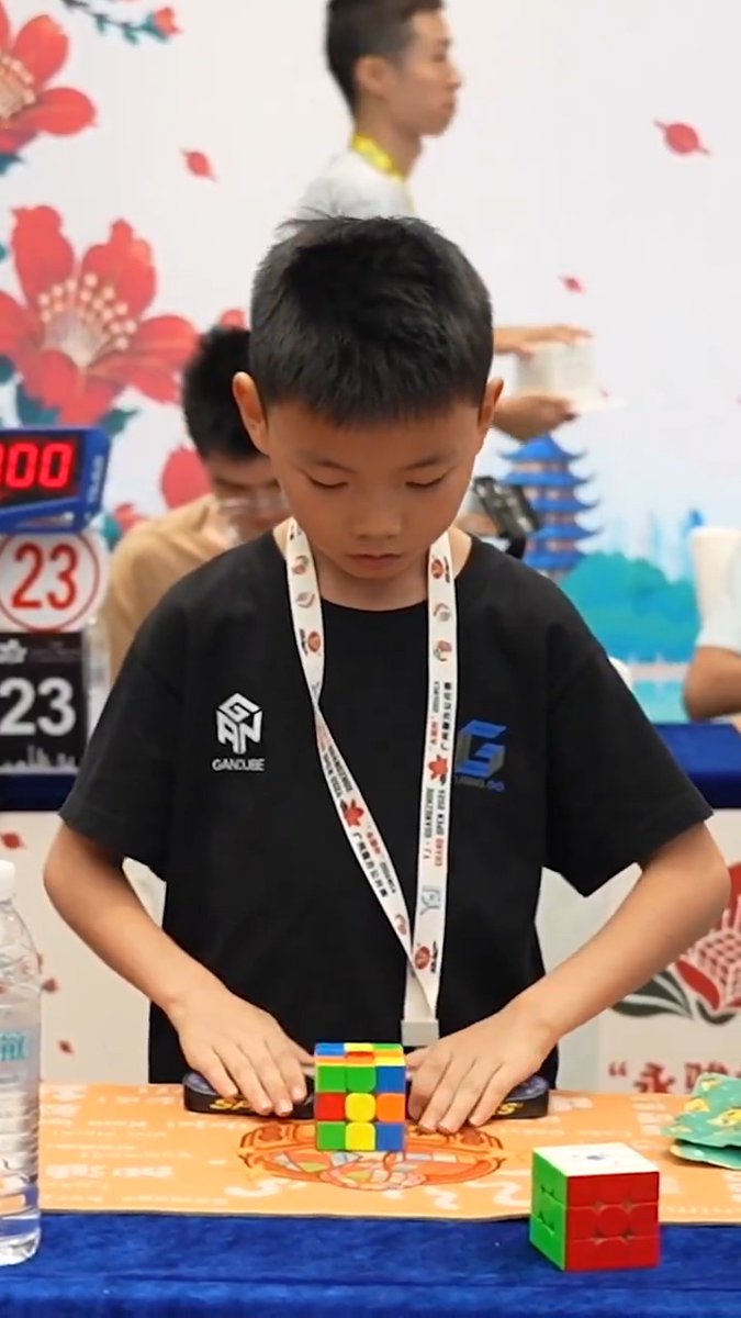 A young boy wearing a black GANcube t-shirt and a lanyard concentrates on solving a Rubik's cube at what appears to be a speedcubing competition.