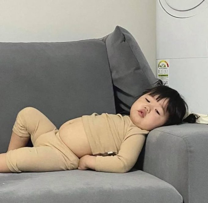 A young child with dark hair rests against a gray sofa, wearing a beige outfit while looking at the camera with a calm expression.