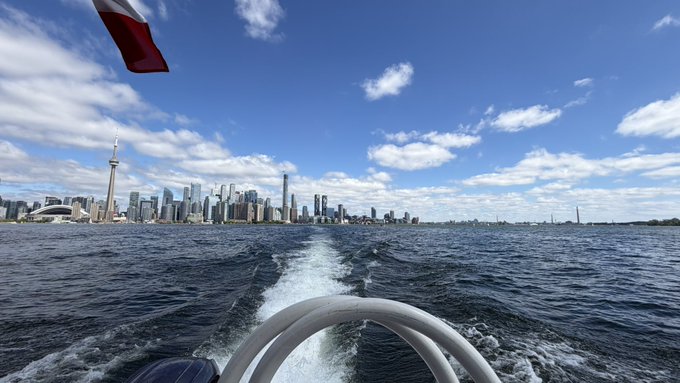 The image shows a boat speeding through the water, leaving behind a large wake that stretches out towards the city skyline. The boat's wake appears to be disturbed by the presence of a flag flying from the boat. In the background, there are tall buildings, including one with a spire on top, which is likely a prominent landmark in the city. The image also contains text that reads "been touching grass ( water ) Beautiful view, enjoying it while the market is nuking been also catching up on the @bankrbot is getting bigger and they". This suggests that someone might be taking a break from their daily routine to enjoy the beautiful scenery of the city skyline.
