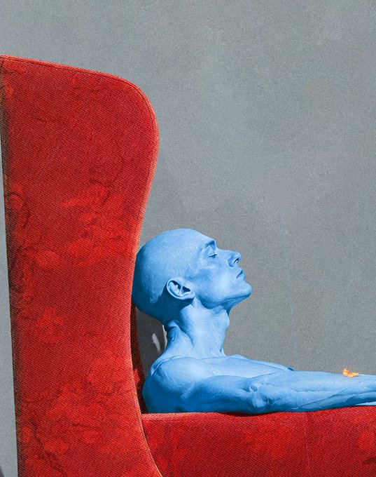 An artistic photograph of a person painted entirely in blue reclining in a large red velvet high-backed chair against a gray wall.