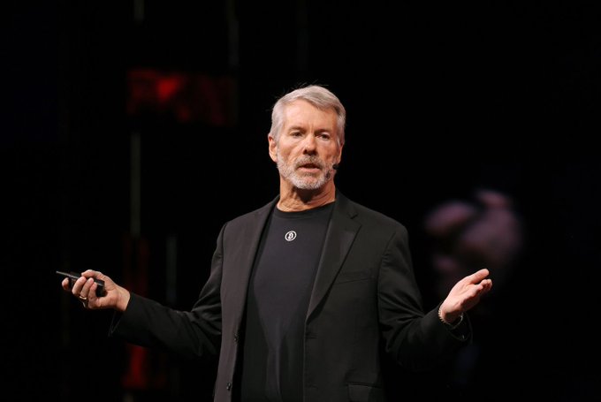 A gray-haired man in a black blazer and dark t-shirt speaks on stage with his arms outstretched, holding a small remote.