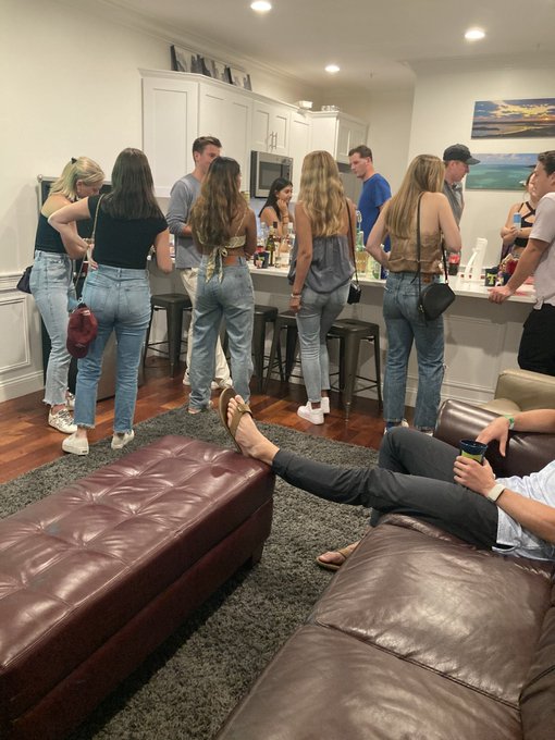 A group of people gathered in a kitchen and living room, with some standing at a counter and others relaxing on a leather couch in the foreground.