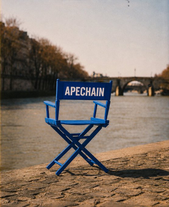 A bright blue director's chair with "APECHAIN" written in white text is positioned on a cobblestone path next to a river, hinting at an exciting event related to the $APE ticker.