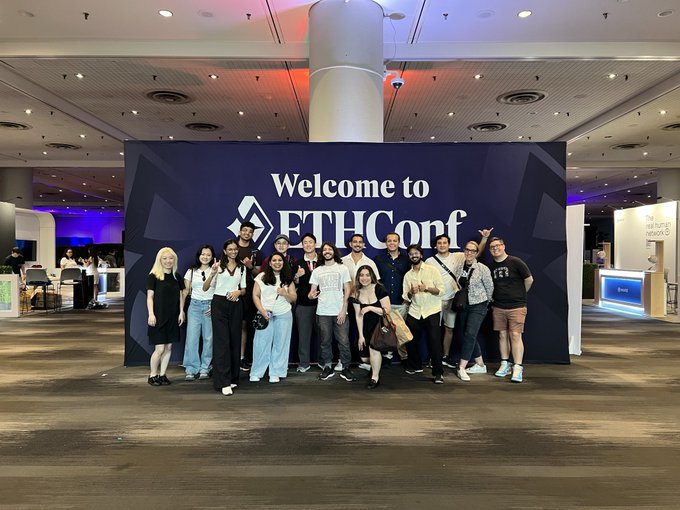 The image shows a group of people standing together, posing for a photo at an event or gathering. They are all wearing casual attire, with some individuals dressed more formally than others. The group consists of both men and women, creating a diverse mix of ages and styles. In front of them is a large sign that reads "Family is locked in for @ethconf", indicating the nature of the event they are attending or participating in.