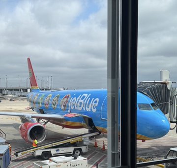 A JetBlue airplane is parked at an airport gate, visible through a terminal window.