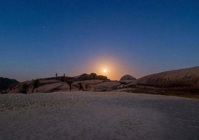 A moonlit night sky over a rocky landscape with people standing on the boulders near a beach.