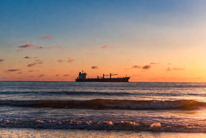 A serene sunset scene over the ocean with a silhouetted cargo ship on the horizon and gentle waves rolling onto the shore under a colorful sky.