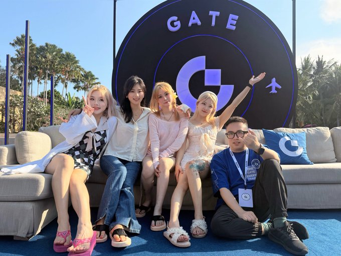 Four young women and one young man posing on a couch in front of a large circular 'GATE' sign at an outdoor event.