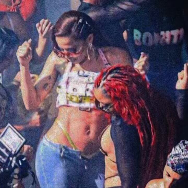 The image shows a woman wearing blue jeans dancing with another person who has red hair. They are both smiling as they dance together. The background of the image features several people, some of whom appear to be taking pictures or recording videos using their cell phones.