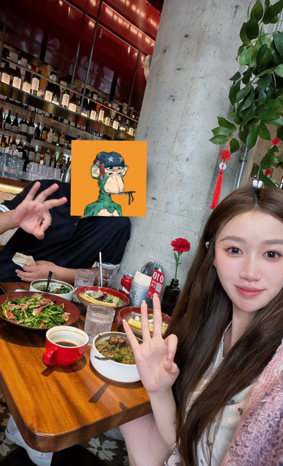 A selfie of a young woman making a peace sign at a restaurant table with various dishes and a drink, while a person next to her has a Bored Ape-style NFT image covering their face.