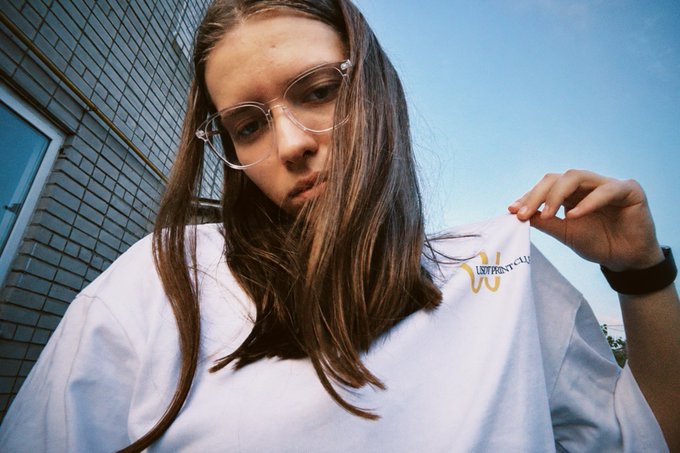 A young woman with long brown hair and clear-framed glasses poses in a white t-shirt featuring a small "USDT PRINT CLUB" logo, mirroring the playful "McDonald's uniform" aesthetic referenced in the post's gm greeting.
