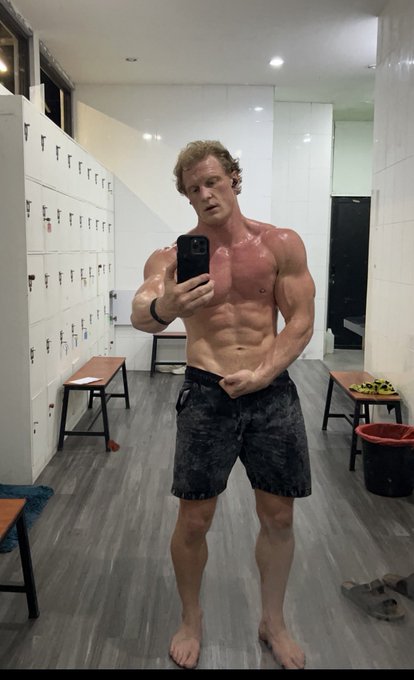 A shirtless muscular man taking a mirror selfie in a locker room, holding a smartphone while wearing dark shorts, surrounded by white lockers and wooden benches.