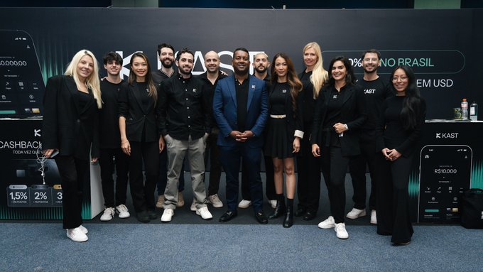 The image shows a group of eleven professionals posing together at a KAST booth, with visible branding including "KAST," "NO BRASIL," "EM USD," and metrics like "1.5%," "2%," and "R$10,000." The setting appears to be the Blockchain Rio event in Brazil, connecting to the post's celebration of Brazil's growing prominence in the stablecoin space.