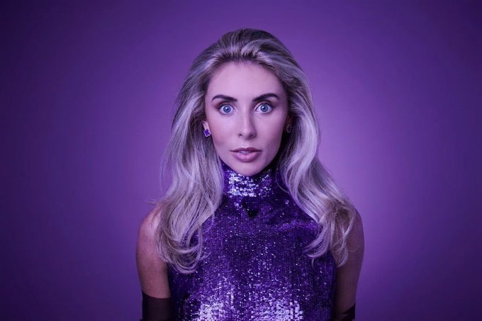 A portrait of a young woman with long, wavy blonde hair and striking blue eyes, wearing a sparkly purple sequined halter top against a solid purple background.