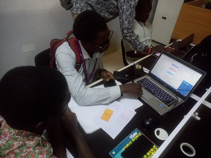 Several people gather around a laptop displaying a software interface, with one man in a checked shirt working with papers and a book titled 'Visio'.