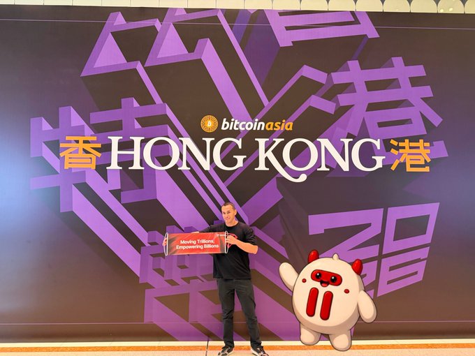 A speaker presents at the Bitcoin Asia 2025 conference in Hong Kong, with the event's purple backdrop displaying "bitcoin asia" and "HONG KONG" alongside Chinese characters, while the TRON mascot stands beside him. The scene captures the TRON team's presence at the event, aligning with the post's excitement about connecting with global blockchain builders.