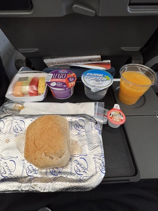 An airline meal tray on a seat-back table features a bread roll, fruit cup, Yoplait yogurt, a small jam container, orange juice, and a coffee cup.