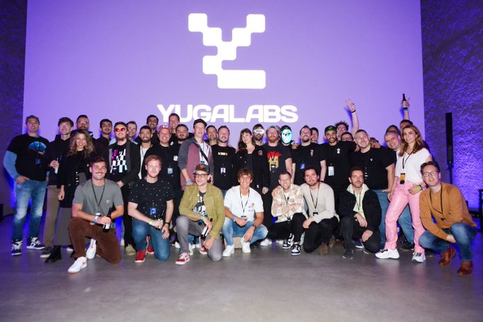 A large group photo of people posing together at a Yuga Labs event, standing in front of a purple backdrop displaying the Yuga Labs logo.