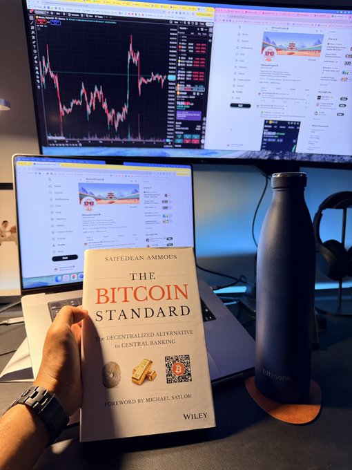 A person holds a copy of "The Bitcoin Standard: The Decentralized Alternative to Central Banking" by Saifedean Ammous, with a foreword by Michael Saylor (Wiley), in front of a laptop and monitor displaying crypto trading charts—reinforcing the post's recommendation of the book as essential reading for anyone in crypto.