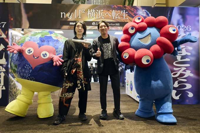 Two men pose with colorful mascots (one blue with eyes, one red with multiple eyes) at an event with a backdrop reading 'null² 横浜へ転生' and '2026'.