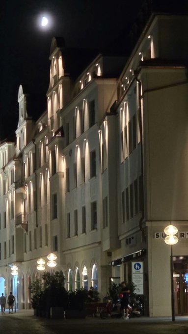 A nighttime photograph of an ornate, illuminated historic building along a city street under a full moon, with a 'STAR' sign visible on a storefront.