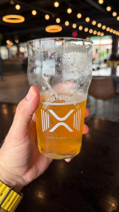 A hand holding a partially filled pint glass of beer from "Oddstory Brewing Co." in Chattanooga, with string lights and a bar visible in the background.
