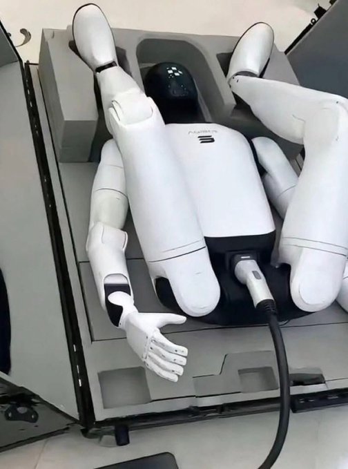 A white humanoid robot lies on its back inside a cardboard box, with its arms bent at the elbows and legs raised, appearing to be packed for shipping.