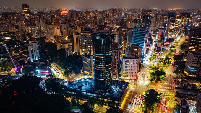 An aerial nighttime view of a sprawling city with illuminated skyscrapers, busy streets, and traffic lights.