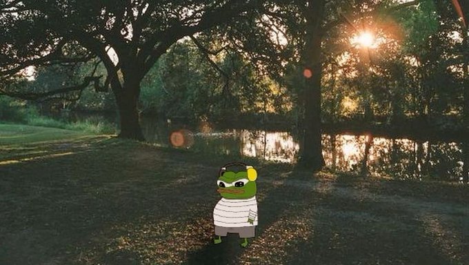 A Pepe the Frog character wearing headphones and a white striped shirt stands on a grassy path beside a calm river at sunset, with golden light filtering through large trees.