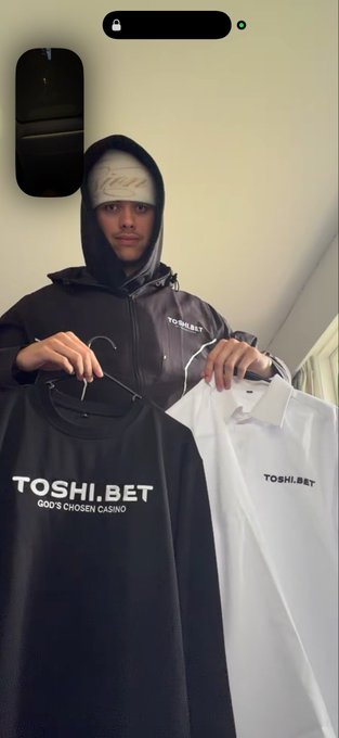 A man wearing a black Toshi.Bet hooded jacket and a white beanie holds up two Toshi.Bet t-shirts—one black reading "TOSHI.BET GOD'S CHOSEN CASINO" and one white with the smaller "TOSHI.BET" logo—showcasing the brand's apparel line that fits the "drip me out in toshi bet" post.