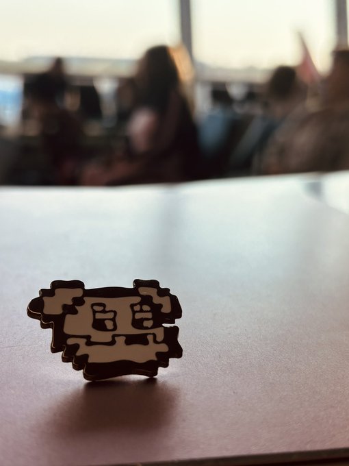 A small, pixel-art style cutout figure resembling a retro video game character standing on a white table, with blurred figures in the background.