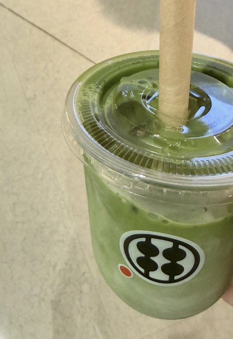 A plastic cup with a domed lid and paper straw contains a green iced beverage, likely a matcha latte. The cup features a distinctive logo resembling a stylized face with black "eyes" and a red dot, though no readable text is visible.